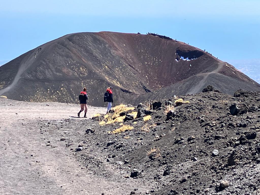The image show two people hiking Etna and walk towards a huge crater - photo taken by Geo Etna Explorer - Etna Guided Tours